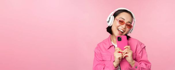 Menina asiática elegante em fones de ouvido, ouvir música e tirar fotos no telefone celular, usando smartphone, de pé sobre fundo rosa - Foto, Imagem