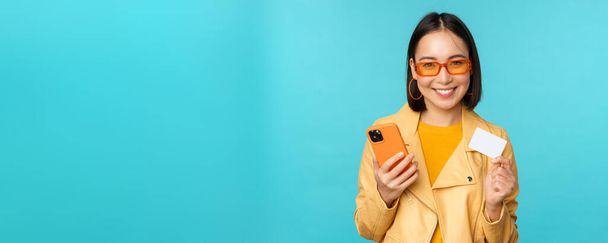 Compras online. Elegante jovem mulher asiática em óculos de sol, mostrando cartão de crédito e usando smartphone, pagando na internet, fazendo compra, de pé sobre fundo azul - Foto, Imagem