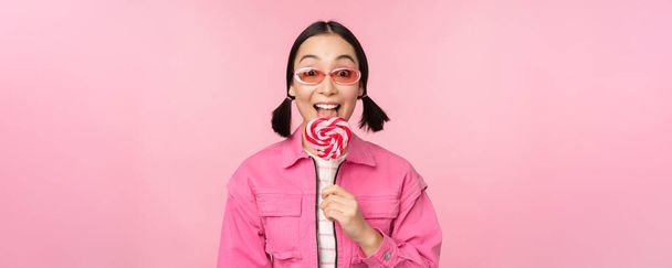 Стильна корейська дівчина облизує льодяник, їсть цукерки та посміхається, стоїть в сонцезахисних окулярах на рожевому фоні
 - Фото, зображення