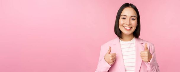 Профессиональная деловая женщина, азиатская корпоративная женщина, показывающая большие пальцы и улыбающаяся, похвала и комплимент, стоящая в костюме на розовом фоне - Фото, изображение