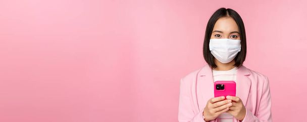 Geschäftsleute und covid-19-Konzept. Japanische Bürodame in Anzug und medizinischer Gesichtsmaske, mit Handy und lächelnd in die Kamera, rosa Hintergrund - Foto, Bild