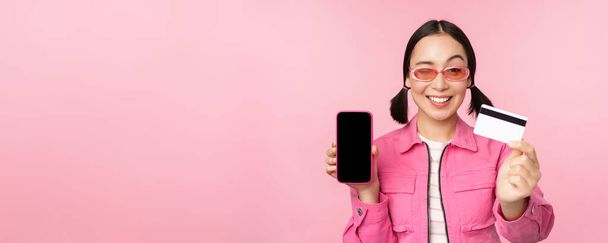 Изображение улыбающейся корейской женщины, показывающей кредитную карту и экран мобильного телефона, интерфейс приложения смартфона, оплату онлайн, покупку бесконтактных, стоя на розовом фоне - Фото, изображение
