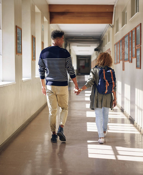 Odprowadza ją na każde zajęcia. Zdjęcie z tyłu kochającej się młodej pary idącej ręka w rękę przez ich korytarz uniwersytecki. - Zdjęcie, obraz