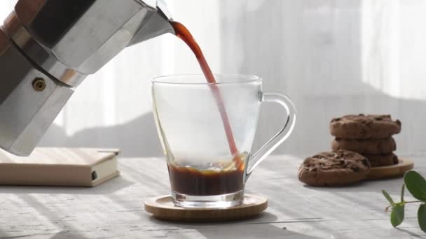 Čerstvá káva se nalévá z moka do průhledného šálku kávy, vedle něj jsou čokoládové sušenky s ranními stíny a ranními paprsky slunce oknem na šedém pozadí stolu - Záběry, video