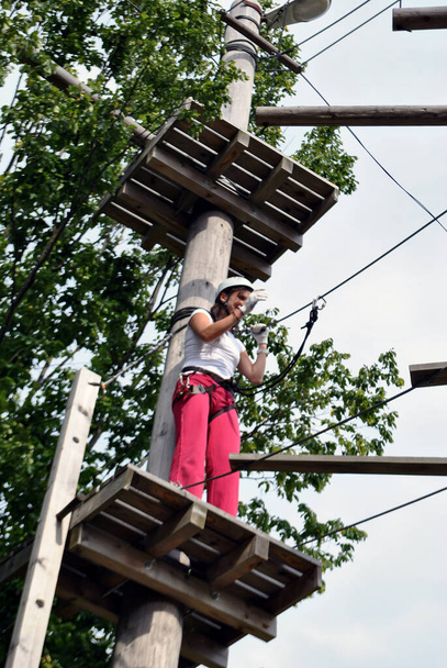 Une jeune femme dans un parc d'attractions se prépare à traverser un pont de corde entre les arbres dans un harnais - Photo, image