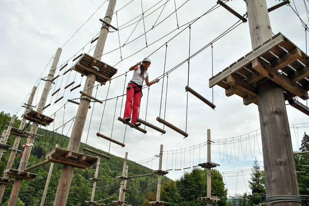 Una giovane donna in un parco divertimenti di corda attraversa un ponte di legno sospeso in un'imbracatura - Foto, immagini