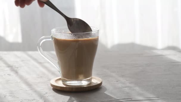 Cukr se nalévá z lžíce do průhledného šálku kávy s mlékem, ranní světlo. Přidání cukru do šálku kávy, lžička je míchání cukru v hrnku - Záběry, video