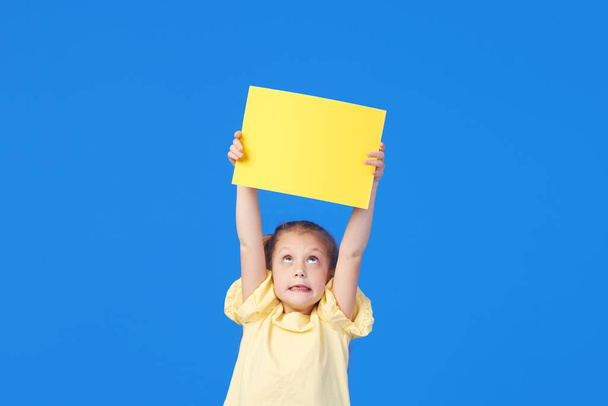A cute little girl is holding a yellow sign for an inscription on a blue background. Funny face. - Photo, image