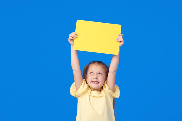 A cute little girl is holding a yellow sign for an inscription on a blue background. Funny face. - 写真・画像