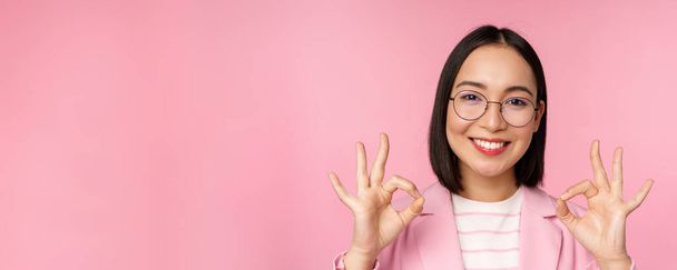 Close up portret imponującej kobiety korporacyjnej, azjatyckiej bizneswoman w okularach, pokazując znak okay, patrząc zdumiony aparatem, polecam, różowe tło - Zdjęcie, obraz