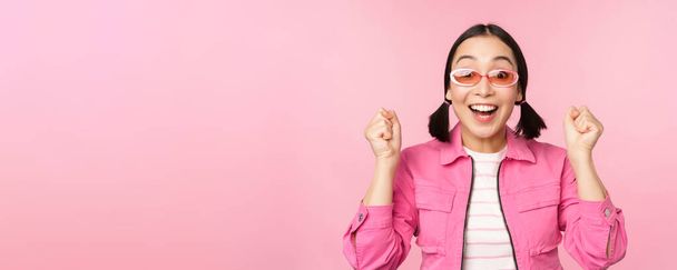 Portrait of excited japanese girl in sunglasses, celebrating, achieve goal, gasping amazed and smiling, standing over pink background - Photo, Image