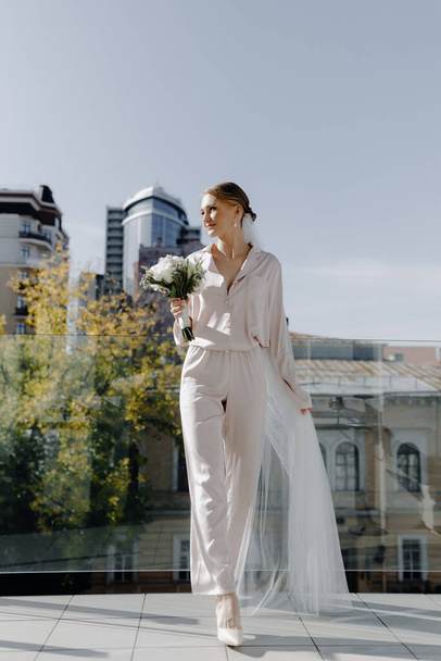  Νύφη με πιτζάμες με ένα μπουκέτο λευκά τριαντάφυλλα στο μπαλκόνι κοιτάζει μακριά. Υψηλής ποιότητας φωτογραφία - Φωτογραφία, εικόνα