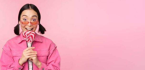 dwaas en schattig aziatische vrouw model likken lolipop, eten snoep zoet en glimlachend, kijken opgewonden, staande over roze achtergrond - Foto, afbeelding