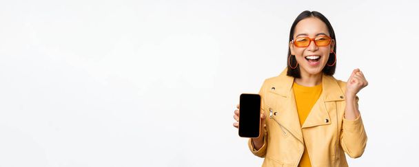 Chica asiática feliz en gafas de sol, mostrando la pantalla del teléfono móvil, interfaz del teléfono inteligente, riendo y sonriendo, celebrando, de pie sobre fondo blanco - Foto, Imagen