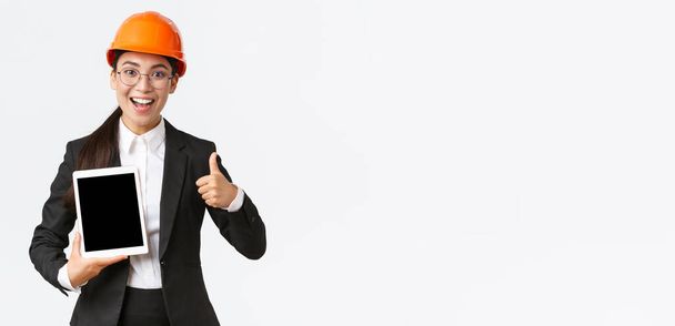 Impressionné jeune agent immobilier montrant bâtiment pendant la construction, porter un casque de sécurité et costume d'affaires, tenant tablette numérique et faire pouce vers le haut, recommander l'achat - Photo, image
