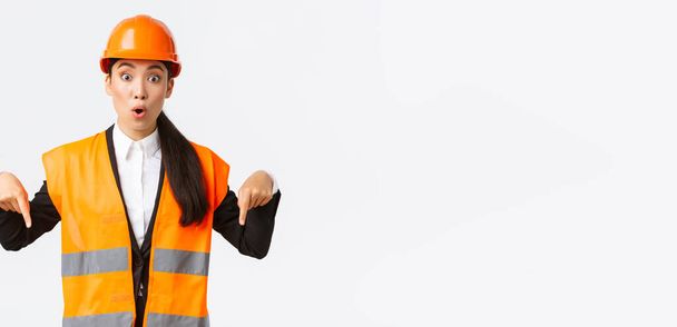 Amazed and interested asian female architect, industrial woman in reflective jacket, safety helmet pointing fingers down and looking impressed, talking about building construction, white background - Fotografie, Obrázek