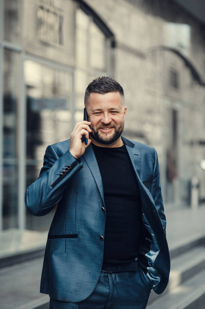 The businessman speaks on phone, on a meeting. The man waves gestures.Attractive handsome young brunet bearded smiling executive worker man in office work station work place, talking on the phone - Фото, изображение