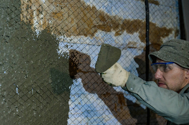 Ο κύριος ρίχνει ένα διάλυμα υγρού σκυροδέματος στον τοίχο, το πρώτο στρώμα σοβά στον τοίχο, μεταλλικό πλέγμα συνδεδεμένο σε έναν ξύλινο τοίχο. - Φωτογραφία, εικόνα