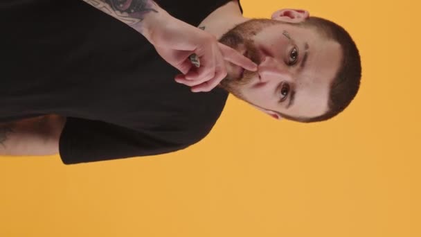atrakcyjny brodaty mężczyzna z tatuażami umieścić palec na jego usta pomarańczowy tło studio strzał średni strzał zawroty głowy wideo - Materiał filmowy, wideo