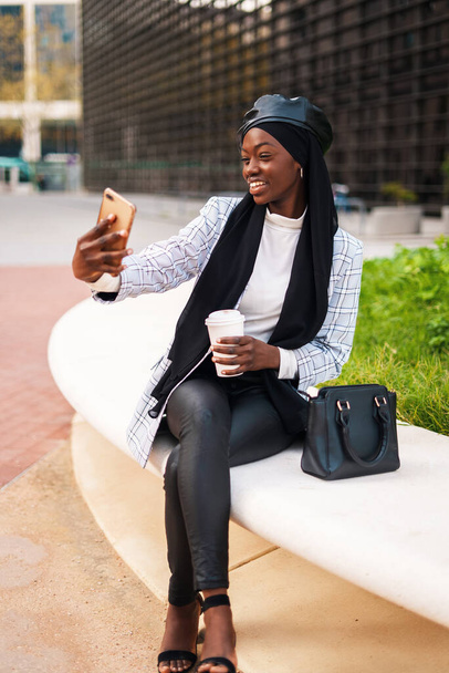 ポジティブスタイリッシュなイスラム教徒黒女性取ります自撮り上のスマートフォン - 写真・画像