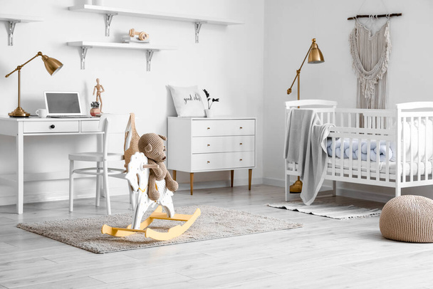 Wnętrze jasnego pokoju dziecięcego z łóżeczkiem, nowoczesnym miejscem pracy i niedźwiedziem zabawką siedzącym na koniu bujanym - Zdjęcie, obraz