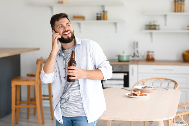 Schöner bärtiger Mann mit Bierflasche im Gespräch per Handy in der Küche - Foto, Bild