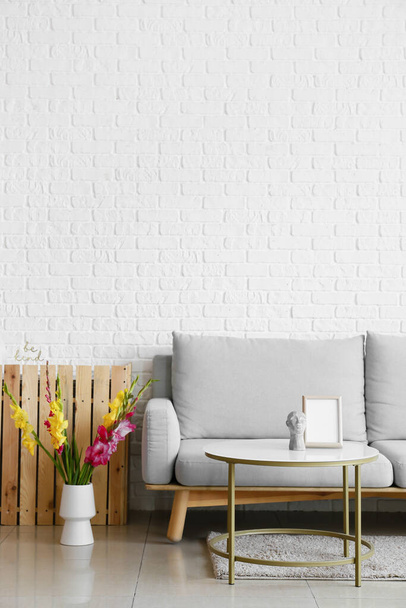 Cómodo sofá y jarrón con hermosas flores de gladiolo cerca de la pared de ladrillo blanco - Foto, imagen