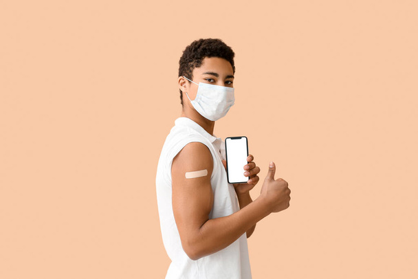Jonge Afro-Amerikaanse man in masker met toegepaste medische patch en mobiele telefoon met duim-up op beige achtergrond - Foto, afbeelding