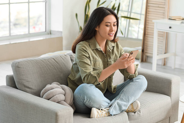 Красивая молодая азиатка с мобильным телефоном сидит дома на диване - Фото, изображение