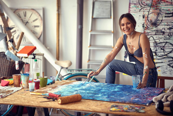 A young sexy and relaxed female artist is posing for a photo in a pleasant atmosphere in her studio. Art, painting, studio - Фото, изображение