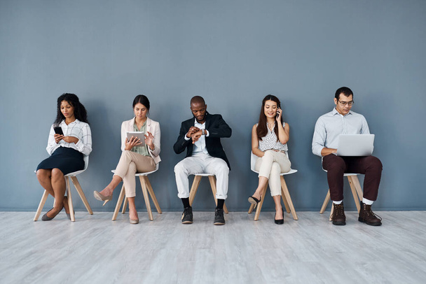 Waiting their turn for a shot at career advancement. Shot of a group of businesspeople using digital devices while sitting in line against a grey background. - Foto, imagen