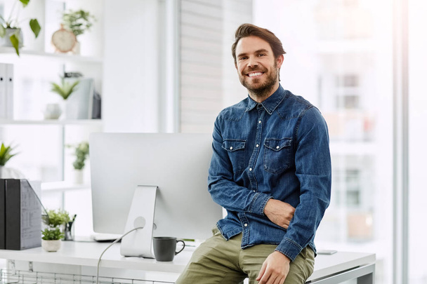 The moment when the office is the best place to be. Cropped portrait of a handsome young businessman smiling while sitting on his desk in a modern office. - 写真・画像