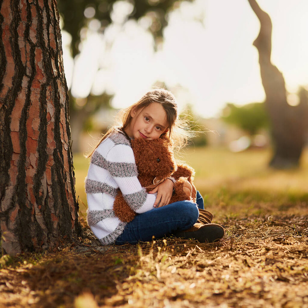 Иногда все, что тебе нужно, это объятия твоего медведя. Портрет милой маленькой девочки, обнимающей своего плюшевого мишку во время игры на улице. - Фото, изображение