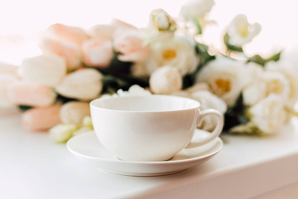 Una tazza bianca e piattino si trovano vicino ai fiori su uno sfondo leggero - Foto, immagini