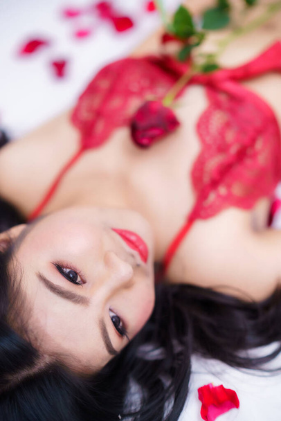 Tanned woman's body,Thailand Asian sexy young woman model in sexy red lingerie holding a red rose posing in bed with rose petals in the bedroom ,valentine's day concept - Photo, Image