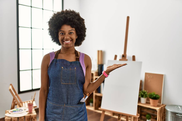 Νεαρή Αφρικάνα Αμερικανίδα με αφρο μαλλιά στο art studio χαμογελώντας χαρούμενα παρουσιάζοντας και δείχνοντας με παλάμη το χέρι κοιτάζοντας την κάμερα.  - Φωτογραφία, εικόνα