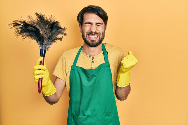Young hispanic man wearing apron holding cleaning duster screaming proud, celebrating victory and success very excited with raised arm  - Photo, Image