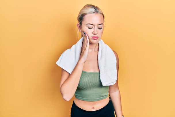 Spor kıyafetleri ve havlu giyen genç sarışın kız diş ağrısı ya da diş hastalığından dolayı acı dolu bir ifadeyle ağıza dokunuyor. dişçi  - Fotoğraf, Görsel