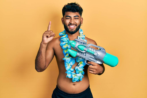 Arab férfi szakállas fürdőruhában és hawaii lei kezében vízipisztollyal mosolygott egy ötlettel vagy kérdéssel ujjal mutogatva boldog arccal.  - Fotó, kép