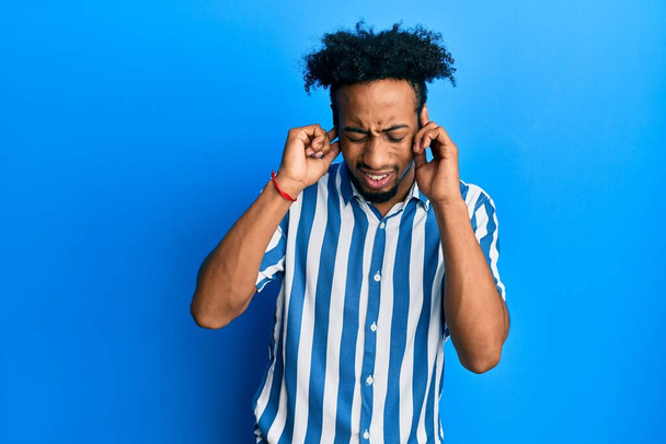 Νεαρός Αφροαμερικάνος με γενειάδα φοράει ριγέ πουκάμισο που καλύπτει τα αυτιά με δάχτυλα με ενοχλημένη έκφραση για το θόρυβο της δυνατής μουσικής. κωφή έννοια.  - Φωτογραφία, εικόνα