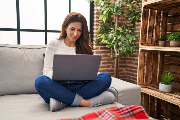 Молода іспаномовна жінка використовує ноутбук, сидячи на дивані вдома
 - Фото, зображення