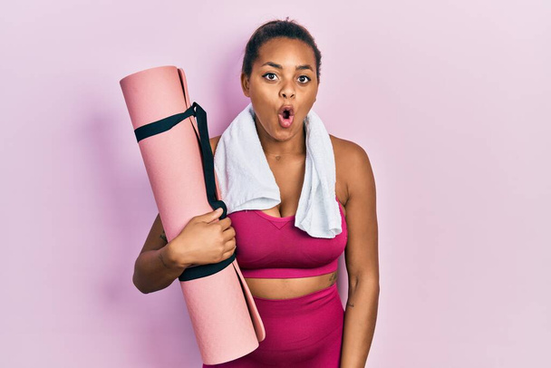 Young african american girl wearing sportswear holding yoga mat scared and amazed with open mouth for surprise, disbelief face  - 写真・画像
