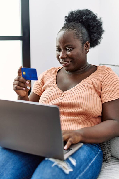 Jeune femme afro-américaine utilisant ordinateur portable et carte de crédit assis sur le canapé à la maison - Photo, image