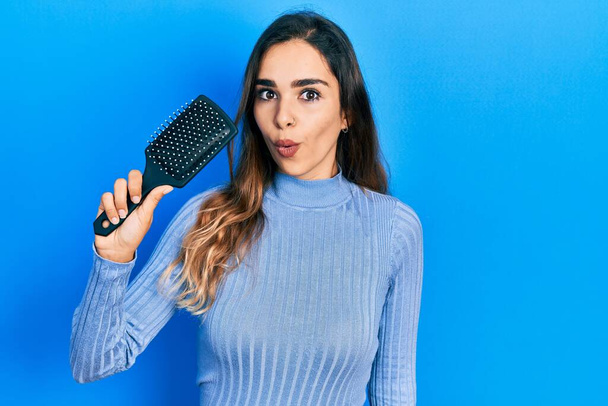 Young hispanic girl holding hairbrush scared and amazed with open mouth for surprise, disbelief face  - Photo, Image