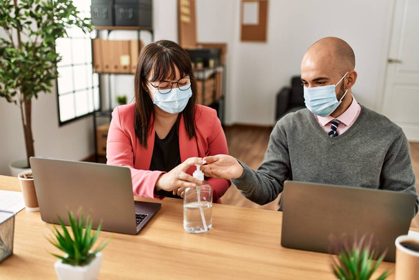 Двое латиноамериканских бизнесменов в медицинской маске используют гель для рук в офисе. - Фото, изображение