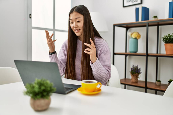 młoda chińska dziewczyna mając wideo rozmowy za pomocą laptopa siedzi na stole w domu. - Zdjęcie, obraz