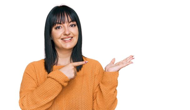 Jonge Spaanse vrouw in casual kleding verbaasd en glimlachend naar de camera terwijl presenteren met de hand en wijzen met de vinger.  - Foto, afbeelding