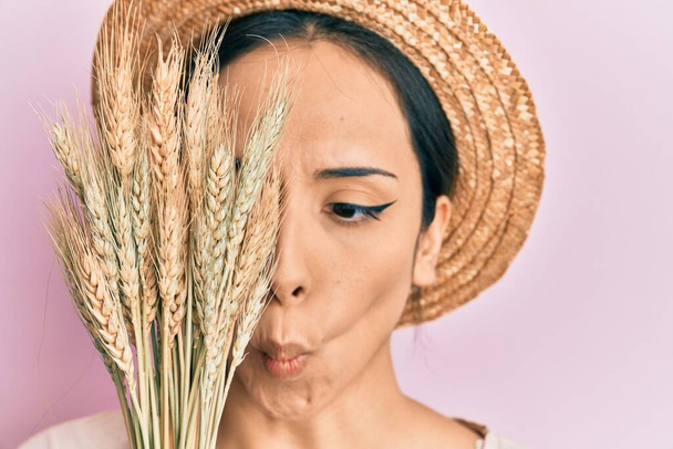 Joven chica hispana con sombrero de verano sosteniendo espiga de trigo sobre el ojo haciendo cara de pez con la boca y los ojos entrecerrados, loco y cómico.  - Foto, imagen