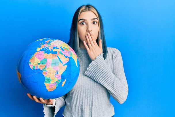 Young modern girl holding world ball covering mouth with hand, shocked and afraid for mistake. surprised expression  - 写真・画像