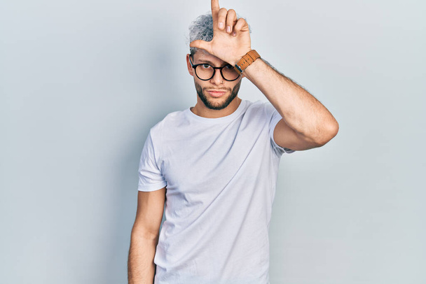 Jonge Latijns-Amerikaanse man met modern geverfd haar in wit t-shirt en bril die mensen met vingers op het voorhoofd voor de gek houdt en losers bespot en beledigt.  - Foto, afbeelding
